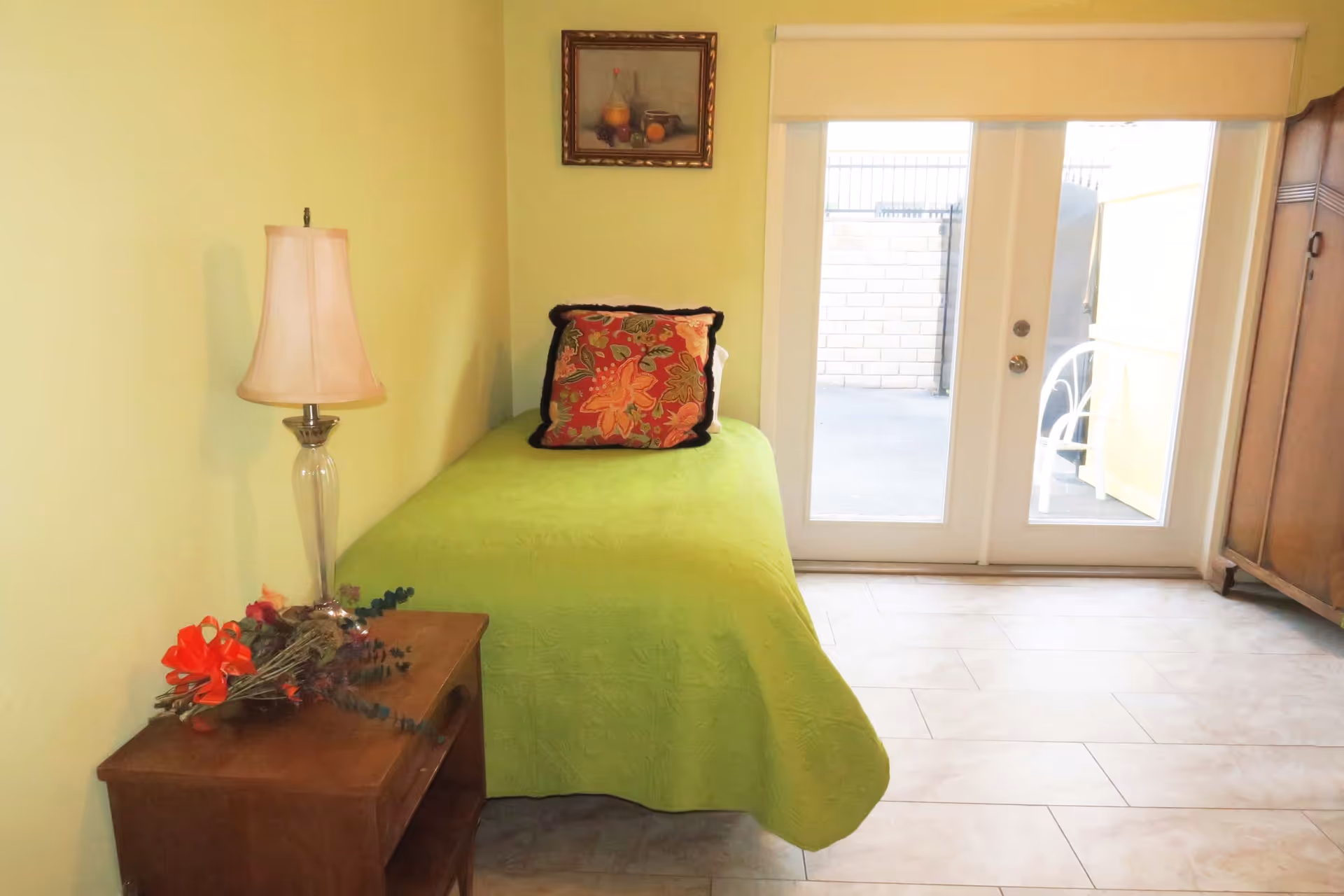 Small bedroom with a single bed covered in a green quilt, a wooden nightstand with a lamp and flowers, and glass doors leading outside.
