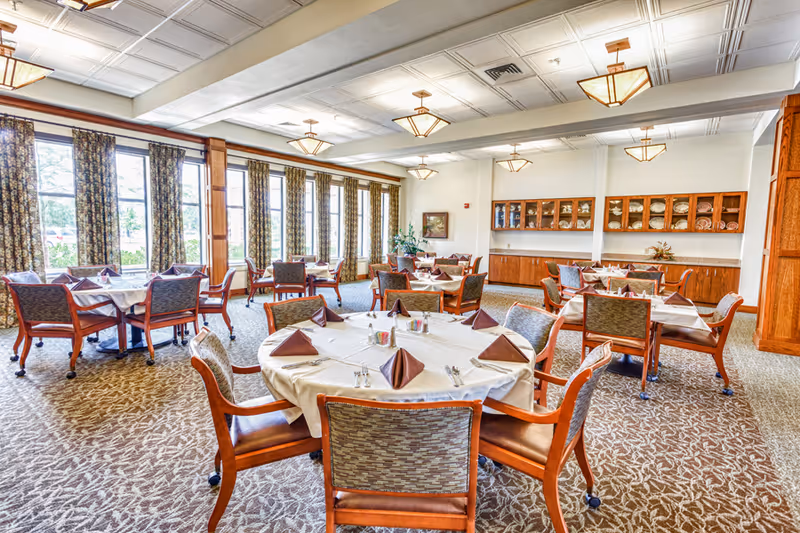 A bright dining room with multiple round and square tables covered with white tablecloths and brown folded napkins. Each table is surrounded by wooden chairs with cushioned seats and backs. Large windows with patterned curtains allow natural light to fill the room. The walls feature wooden cabinetry displaying plates and decorative items. The ceiling has recessed lighting fixtures.
