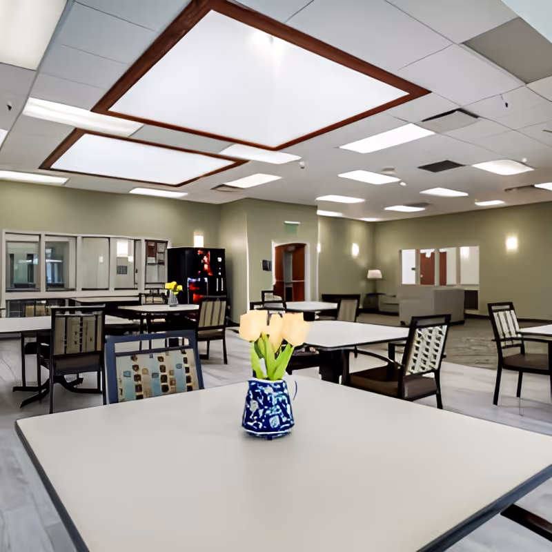A spacious dining area with multiple tables and chairs arranged neatly. A small vase with yellow tulips is placed on the closest table. The room has soft green walls, large ceiling lights, and a vending machine in the background. There is also a seating area with couches and a lamp visible in the back.
