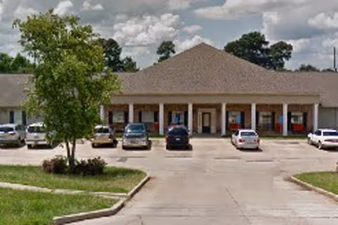 Exterior view of a single-story building with a pitched roof and multiple entrances, several cars parked in front, a tree and some grass in the foreground, under a partly cloudy sky.