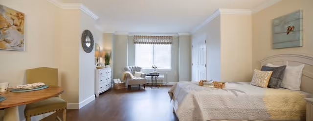 A bright and spacious bedroom in a senior living facility with a large bed covered in a quilt, a small dining table with two chairs, a cushioned armchair near a window with blinds, a white dresser with decorative items, and light-colored walls with artwork.