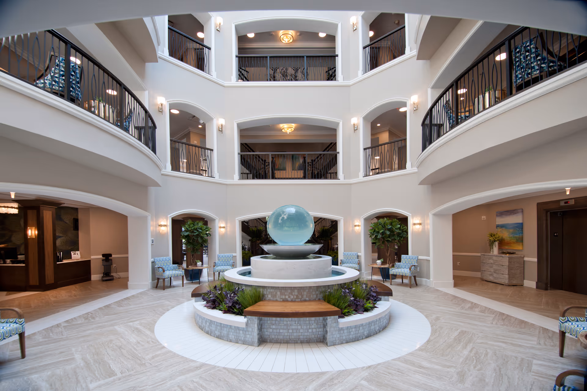 Spacious and elegant multi-level atrium with a central water fountain featuring a large glass sphere, surrounded by circular benches and plants. The area is well-lit with wall sconces and ceiling lights, and has balconies with black railings on the upper floors. Comfortable patterned chairs and potted plants are placed around the space, creating a welcoming atmosphere.
