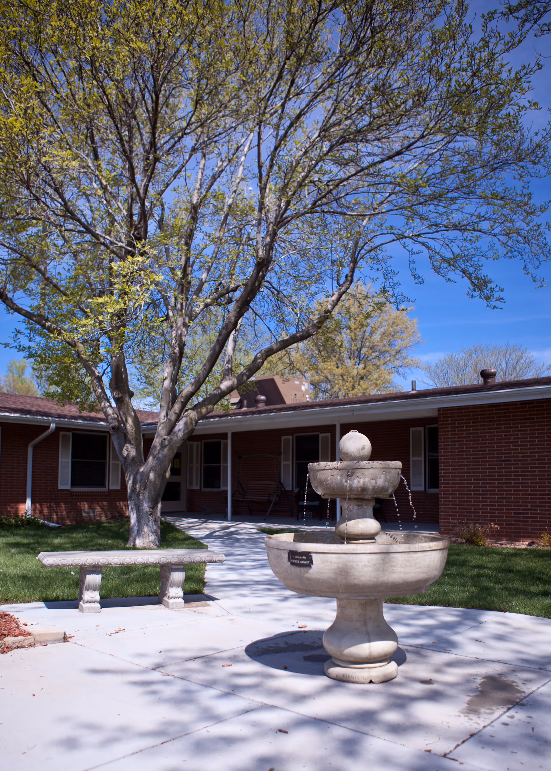 Outdoor courtyard area with a large tree, a stone bench, and a two-tiered stone water fountain in front of a single-story brick building under a clear blue sky.