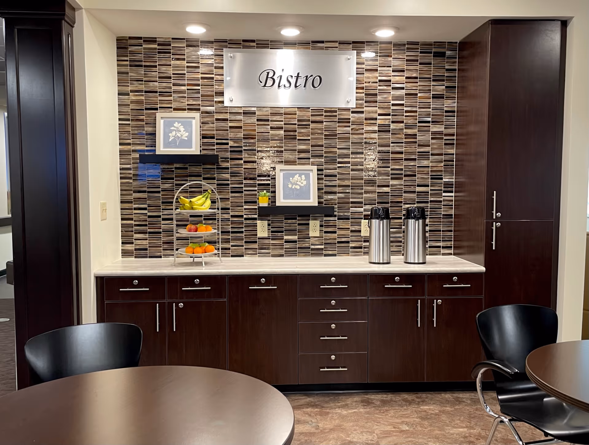 Interior view of a bistro area with dark wood cabinets and a tiled backsplash. There are two stainless steel beverage dispensers on the countertop, a tiered fruit stand with bananas, apples, and oranges, and two framed floral pictures on small black shelves. Two round tables with black chairs are partially visible in the foreground.