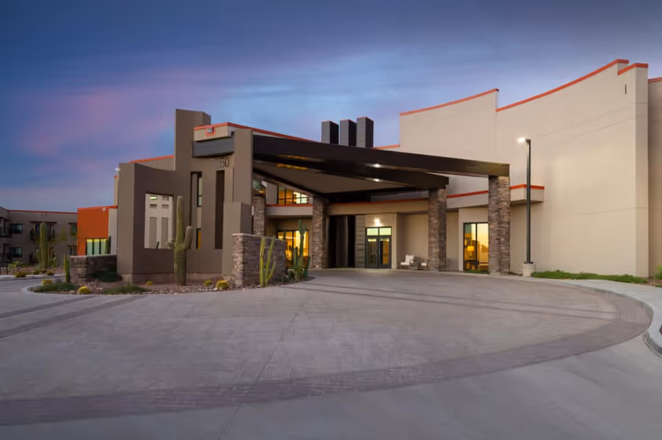 Exterior view of La Sonora at Dove Mountain facility entrance during dusk, featuring modern architecture with stone pillars, a covered driveway, desert landscaping with cacti, and illuminated windows.