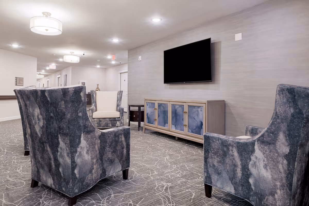 A modern seating area in a hallway with four upholstered armchairs arranged around a wall-mounted flat-screen TV above a wooden cabinet with blue decorative panels. The space features patterned carpet, soft lighting from ceiling fixtures, and light-colored walls.