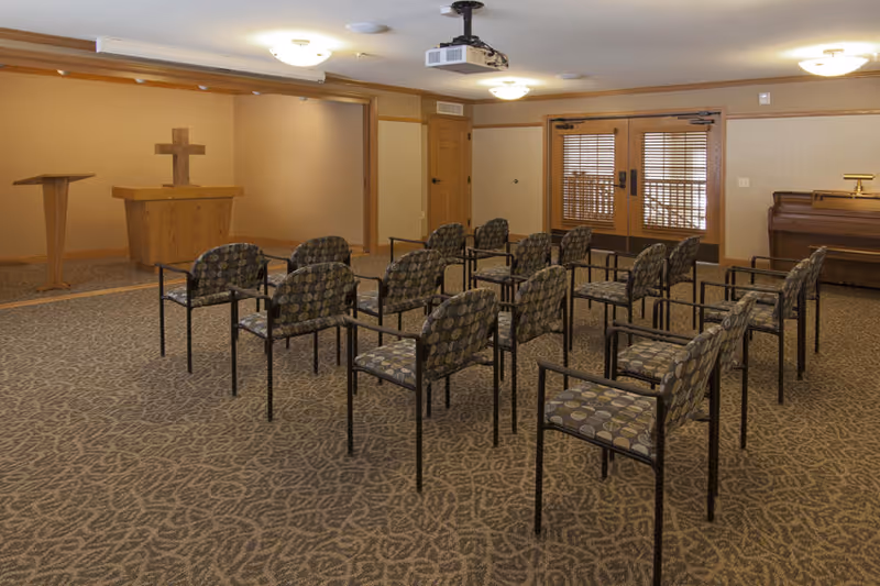 A small chapel or meeting room with rows of patterned chairs facing a wooden podium with a cross on the wall behind it. The room has beige walls, carpeted floor with a leaf pattern, a piano on the right side, and double doors with wooden blinds at the back.