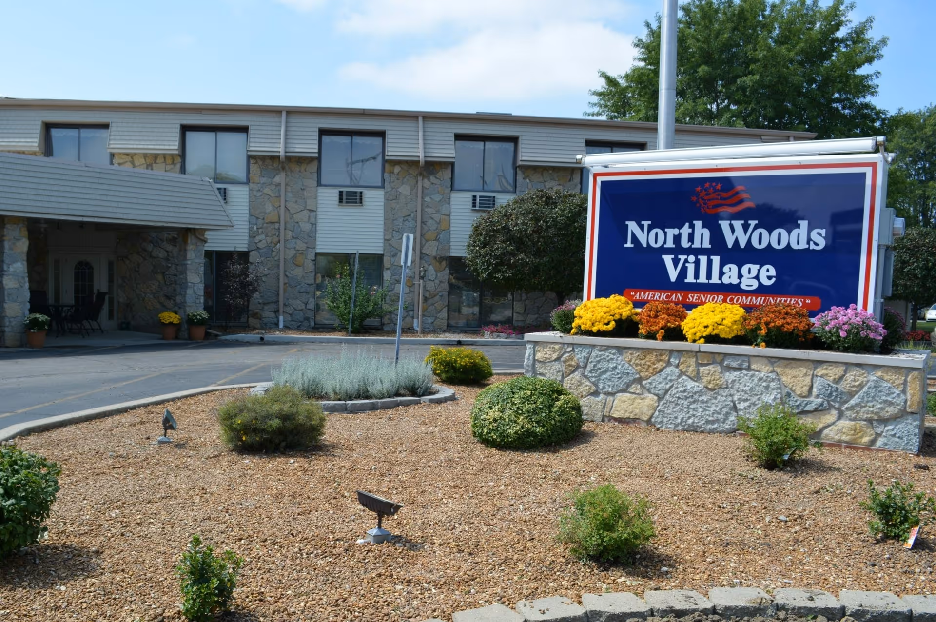 Landscaped entrance and large sign reading 'North Woods Village' in front of a stone-faced senior living building.