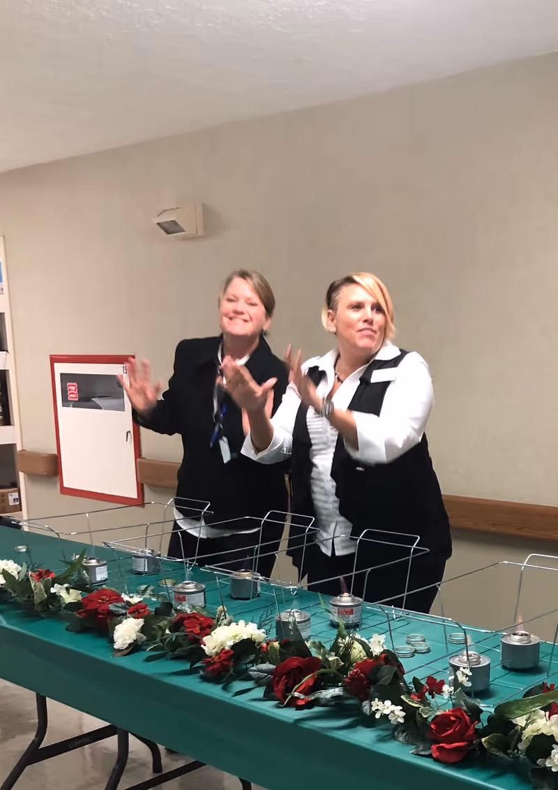 Two women standing behind a table decorated with red and white flowers and several small metal containers with flames. They appear to be clapping or gesturing with their hands in a room with beige walls.