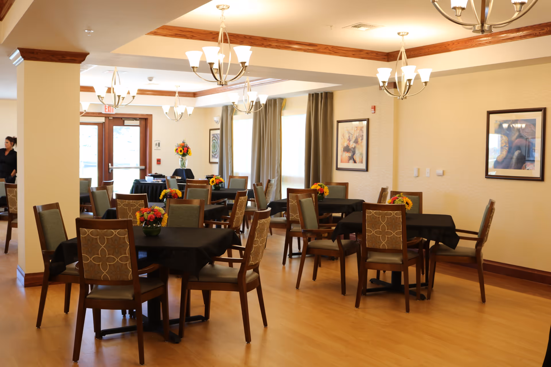 A well-lit dining room with multiple tables covered in black tablecloths, each adorned with small floral centerpieces. The room features wooden chairs with patterned backs, light wood flooring, beige walls with framed artwork, and large windows with curtains. Several modern chandeliers hang from the ceiling, and a woman is partially visible near the entrance door in the background.