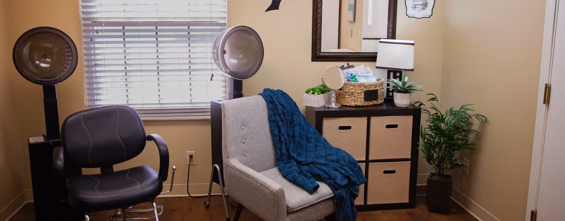 A cozy room with two hair drying stations featuring black salon chairs and hooded hair dryers. A gray armchair with a blue throw blanket is positioned next to a black storage unit with four beige fabric drawers. On top of the storage unit are a small mirror, a basket with magazines, a table lamp, and a potted plant. A large mirror hangs on the wall above the storage unit, and a tall green plant is in the corner near a closed door. The room has beige walls and a window with white blinds.