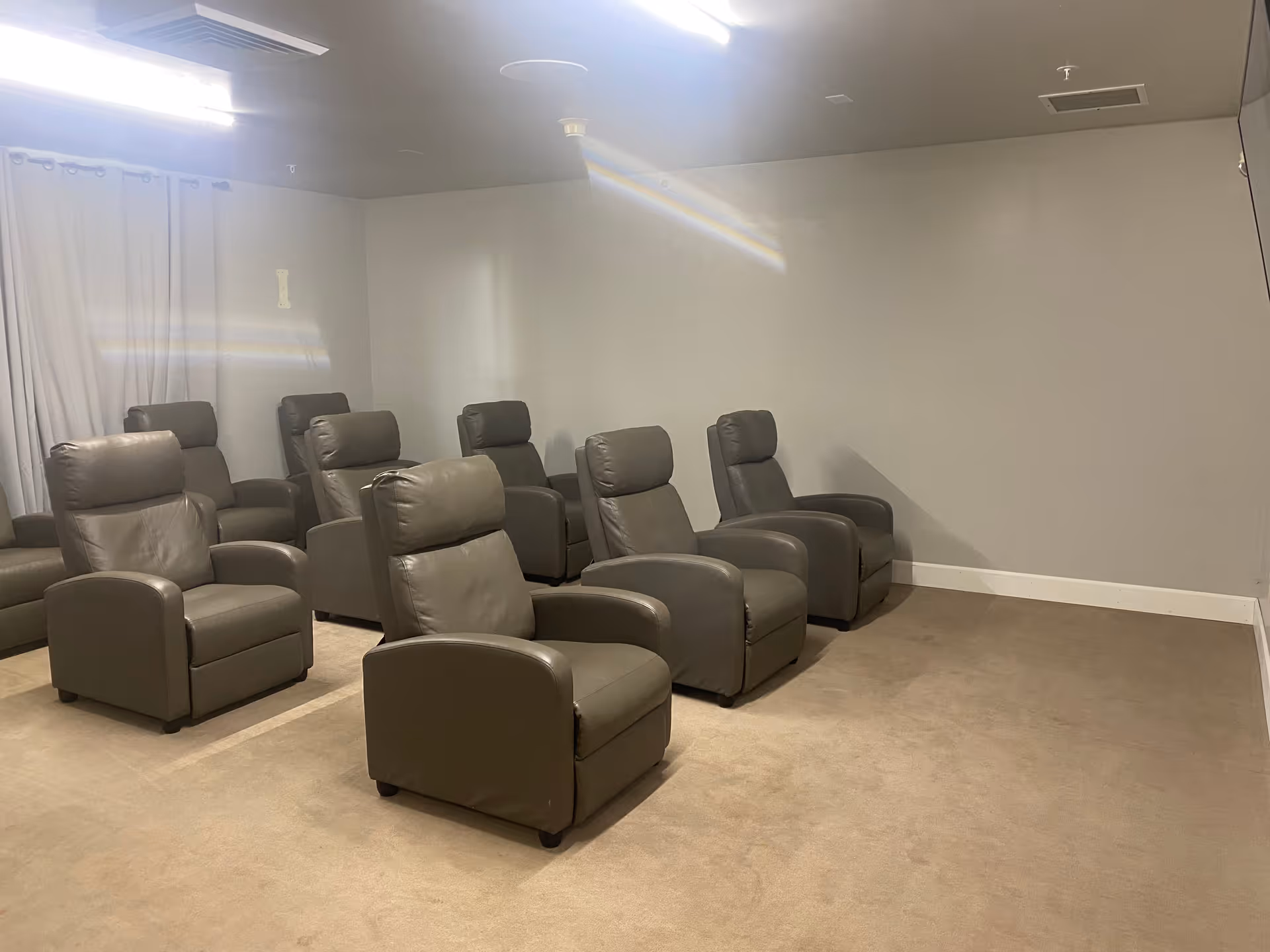 A small room with eight gray leather recliner chairs arranged in two rows facing an empty wall, likely used as a media or presentation room. The room has beige carpet, light gray walls, and a white curtain covering a window on the left side.