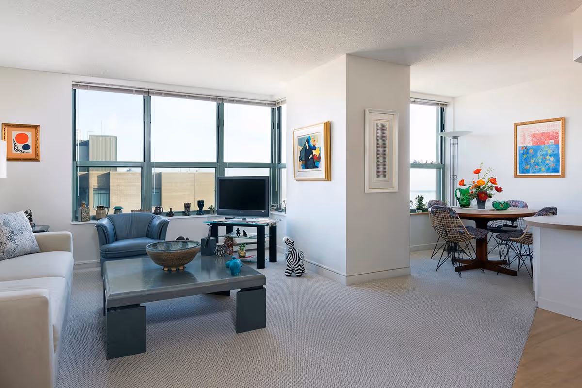 A bright and modern living room area with large windows letting in natural light. The room features a beige sofa with patterned cushions, a blue armchair, a low rectangular coffee table with decorative items, and a TV on a stand near the windows. Adjacent to the living area is a dining space with a round wooden table, four chairs with patterned upholstery, a floor lamp, and colorful artwork on the walls.