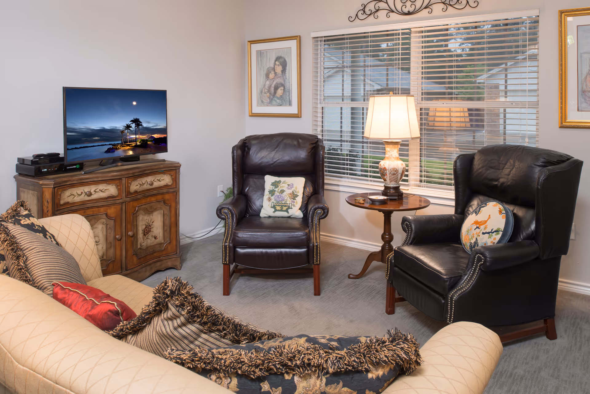 A cozy living room with a beige sofa adorned with multiple decorative pillows, two dark leather armchairs each with a decorative cushion, a wooden side table with a lamp, a window with blinds, and a wooden cabinet holding a flat-screen TV displaying a scenic beach sunset.