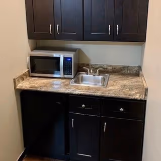 Small kitchenette area with dark wood cabinets, a granite countertop, a stainless steel sink with a faucet, a microwave oven on the counter, and a mini refrigerator below the counter.