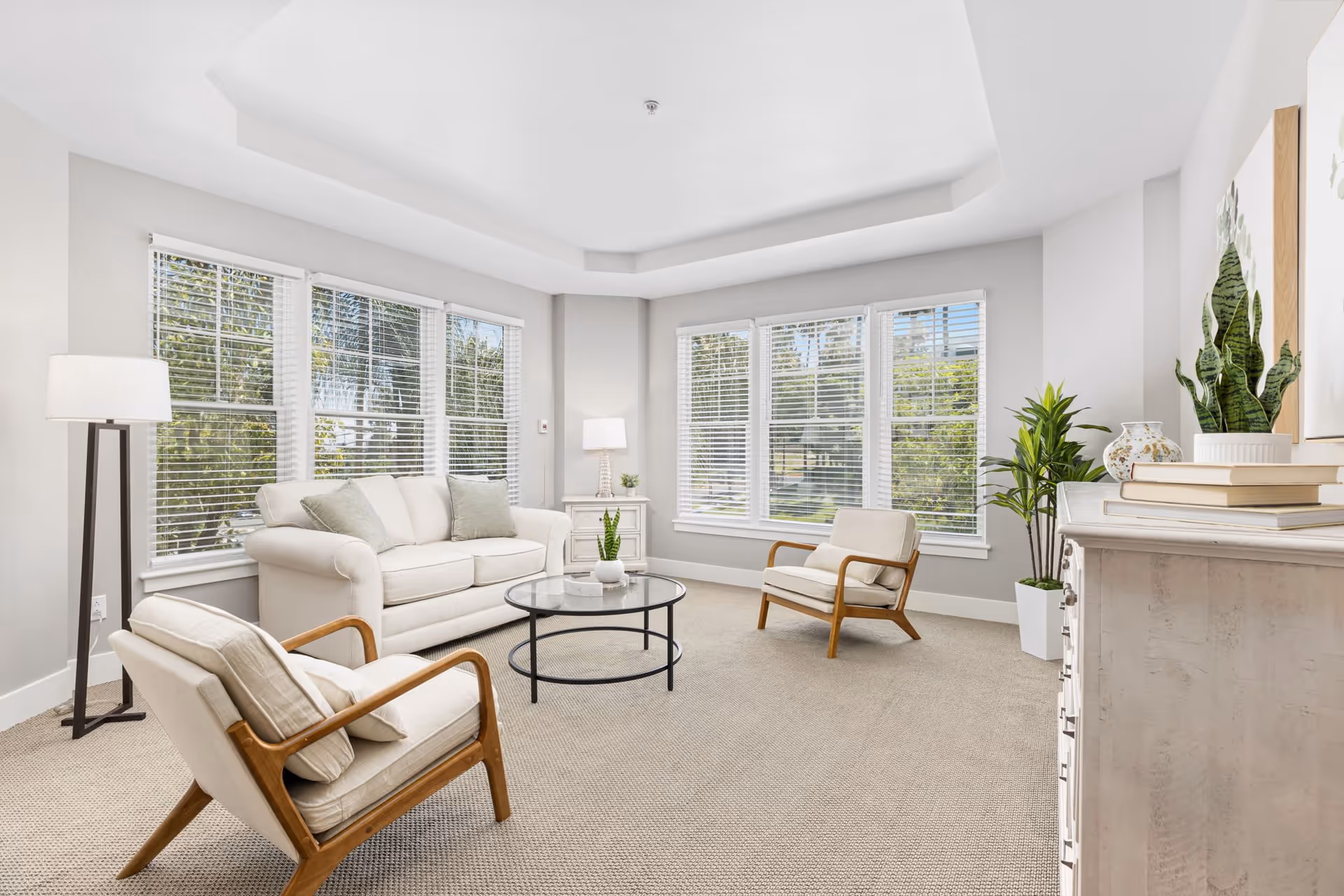 A bright and airy living room with large windows featuring white blinds, a white sofa with cushions, two wooden armchairs with light cushions, a round glass coffee table, a floor lamp, a side table with a lamp, and decorative plants and books on a white dresser.