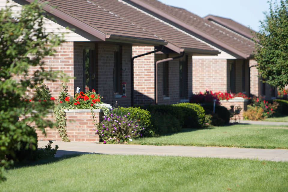 A row of single-story brick buildings with brown shingled roofs, surrounded by green lawns, bushes, and colorful flower beds. A concrete walkway runs in front of the buildings.