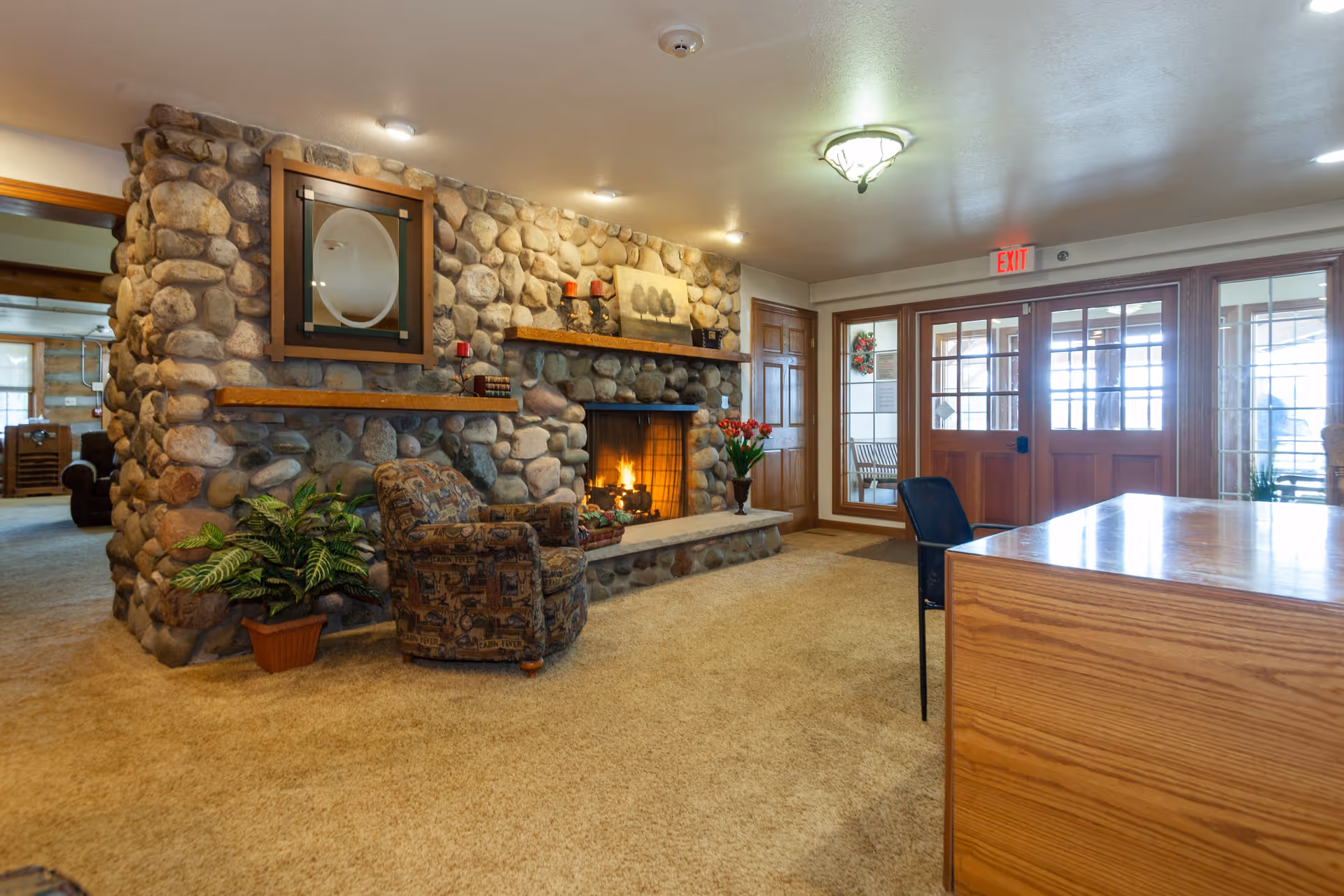 Interior of a senior living facility lounge area featuring a stone fireplace with a fire burning, a patterned armchair, a potted plant, wooden shelves with decorative items, and a wooden desk with a black chair. The room has beige carpet, wooden doors with glass panels, and an exit sign above the door.