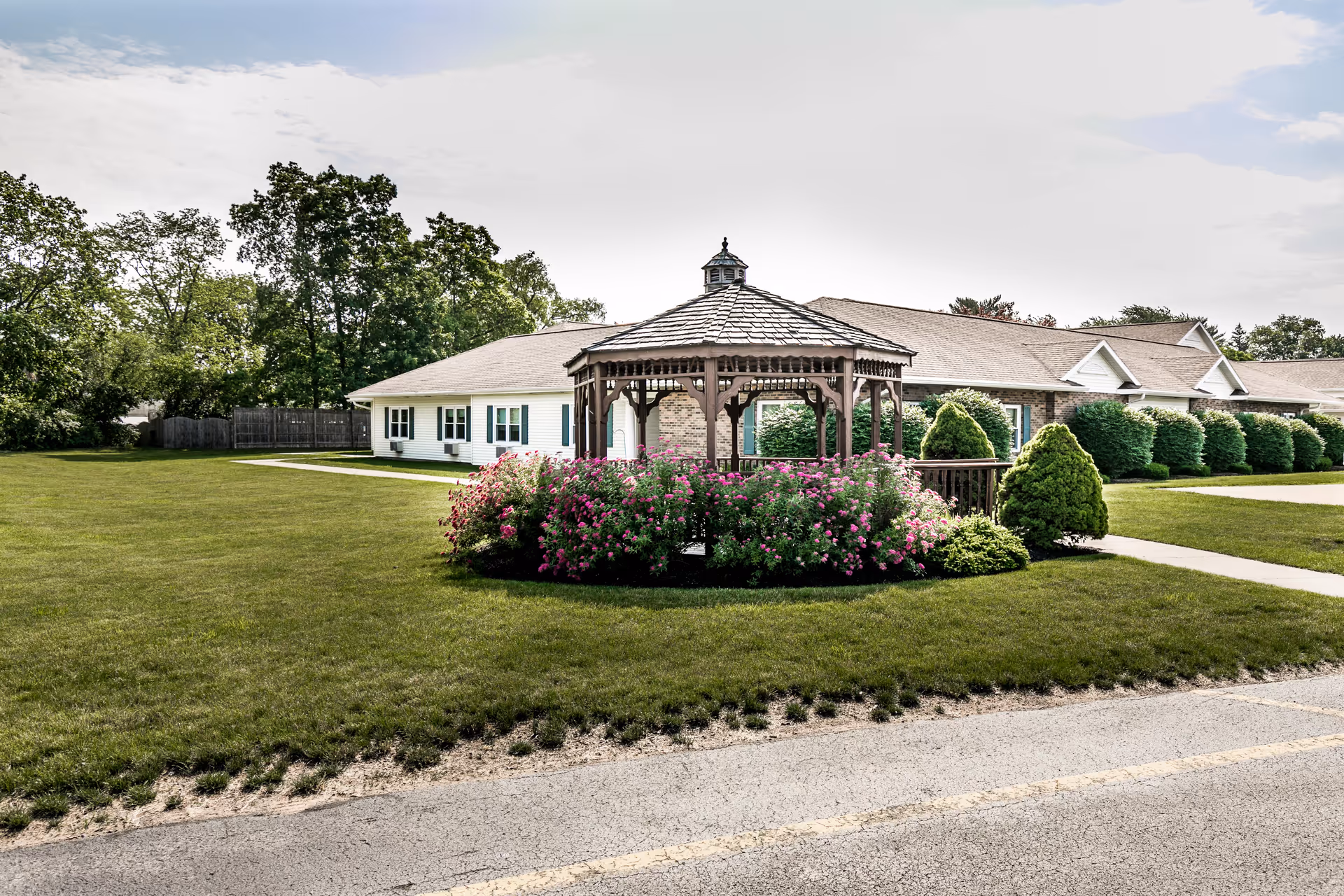 A wooden gazebo surrounded by pink flowering bushes and green shrubs sits on a well-maintained lawn in front of a single-story building with white siding and multiple windows. Trees and a partly cloudy sky are visible in the background.