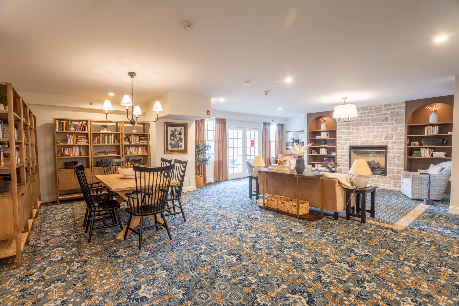 A spacious, well-lit common area featuring a dining table with six black chairs on the left side, wooden bookshelves filled with books behind it, and a cozy living room setup on the right with a sofa, armchair, side tables with lamps, and a stone fireplace flanked by built-in shelves. The room has patterned carpet flooring and large windows with curtains allowing natural light.