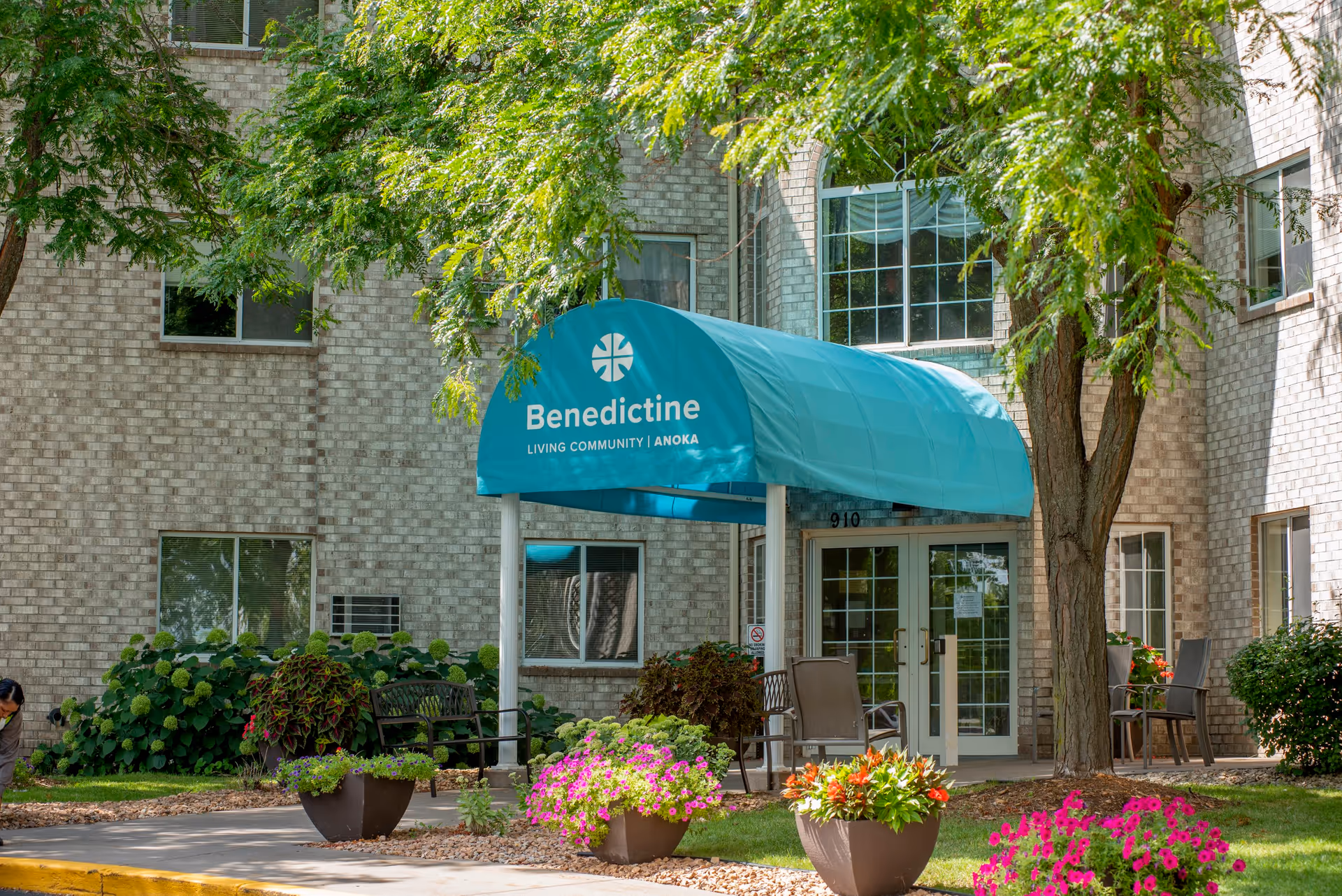 Entrance of Benedictine Living Community-Anoka with a teal awning displaying the facility name, surrounded by trees, potted flowers, and outdoor seating near a brick building.