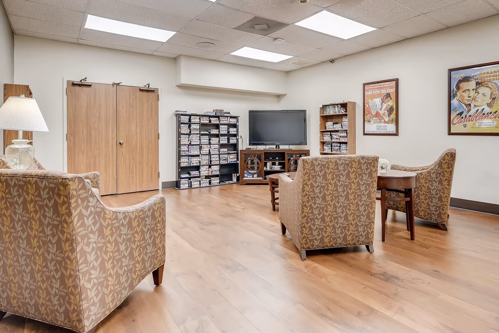 A cozy senior living common room with patterned armchairs arranged around a small wooden table. A flat-screen TV is mounted on a wooden stand against the wall, flanked by shelves filled with DVDs. The room has light-colored walls, wood flooring, and two classic movie posters on the wall. A table lamp is visible on the left side near one of the armchairs.