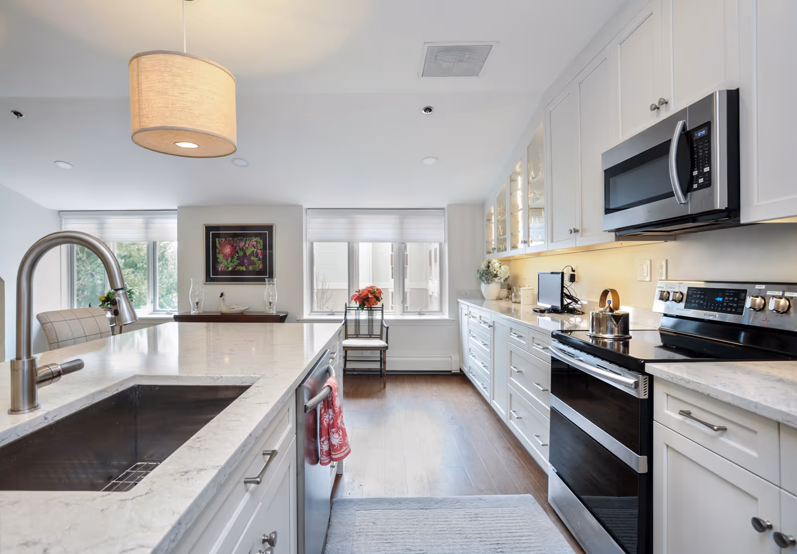 Bright modern kitchen with white cabinetry, stainless steel appliances including a microwave and stove, a large island with a sink and faucet, and a hanging pendant light. There are two windows at the far end letting in natural light, a small table with a flower pot, and wooden flooring.