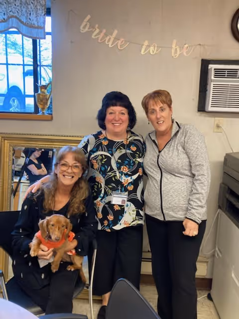 Three women smiling indoors, with one seated holding a small brown dog wearing a red outfit. Behind them is a wall with a banner that reads 'bride to be', a window, and an air conditioning unit.