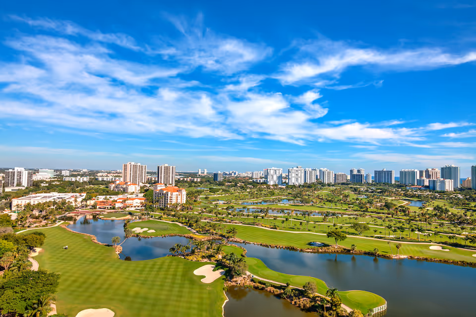 Aerial view of a golf course with green fairways, sand bunkers, and water hazards, surrounded by residential buildings and high-rise apartments under a blue sky with scattered clouds.