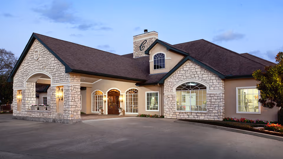 Stone-clad senior living facility front with a covered porte-cochère, arched windows, and illuminated entrance at dusk.
