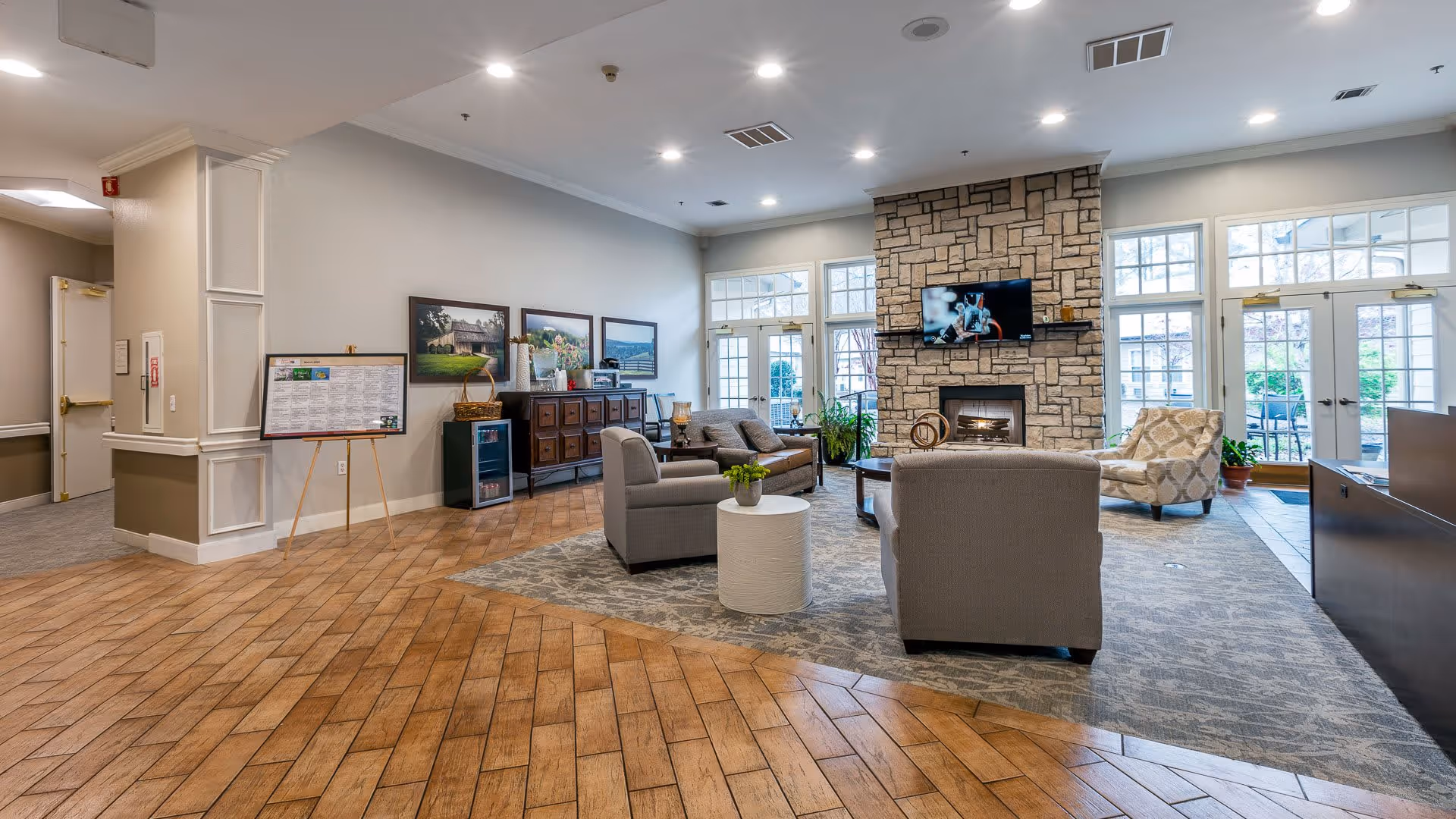 A spacious and well-lit living room area in an assisted living facility featuring a stone fireplace with a mounted TV above it, several comfortable armchairs and sofas arranged around a coffee table, large windows and glass doors letting in natural light, and decorative artwork on the walls.