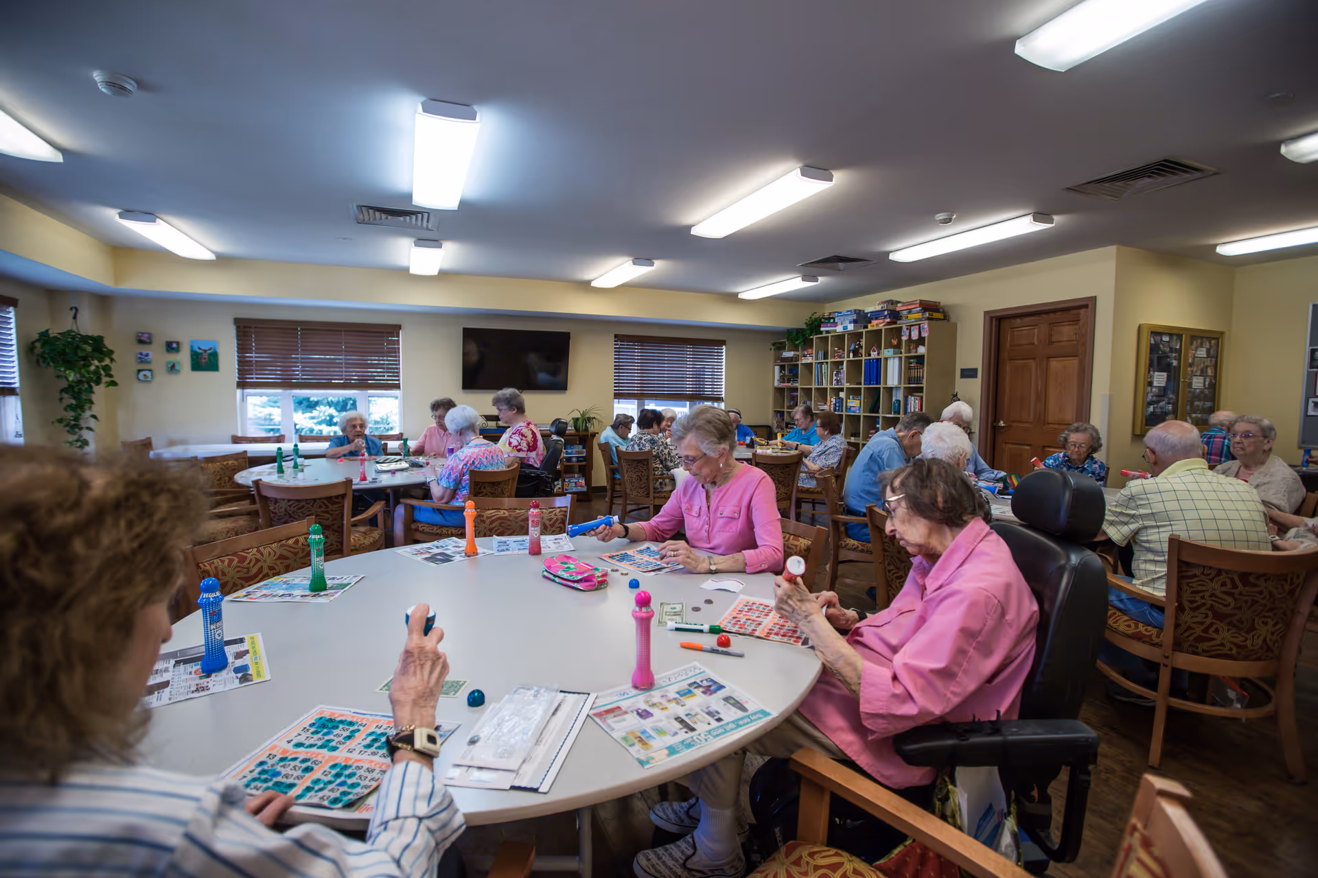 A group of elderly people sitting around several tables in a well-lit room playing bingo. The room has beige walls, wooden chairs with patterned cushions, and shelves filled with board games and books. There are windows with blinds and a television mounted on the wall.