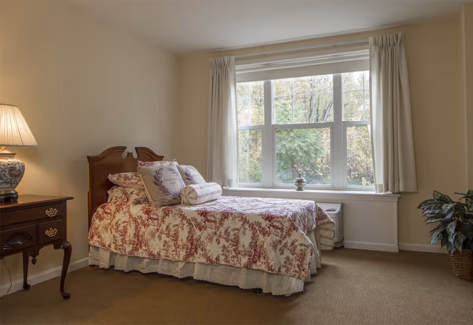 A cozy bedroom with a single bed covered in a red and white patterned quilt and matching pillows. There is a wooden nightstand with a decorative lamp on the left side of the bed. A large window with white curtains lets in natural light, and a potted plant is placed on the floor to the right.
