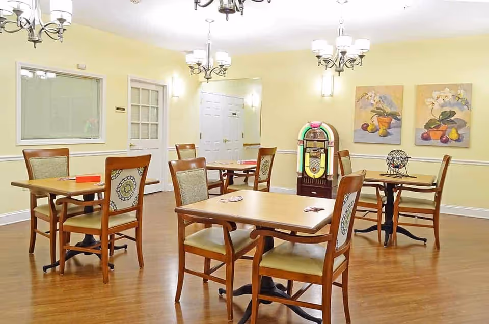 A well-lit room with wooden tables and chairs arranged for small groups. The walls are painted light yellow and decorated with two paintings of flowers and fruit. A vintage-style jukebox stands against the wall, and a bingo cage is placed on one of the tables. The floor is wooden, and multiple chandeliers hang from the ceiling.