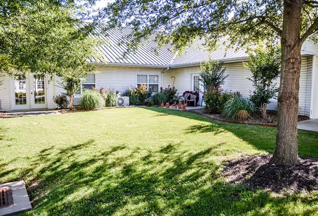 A sunny outdoor courtyard area at The Bungalows at Springdale featuring a well-maintained green lawn, a large tree casting shadows, and a white building with multiple windows and a metal roof. There are shrubs and potted plants along the building's edge, and a small seating area with chairs near a door.