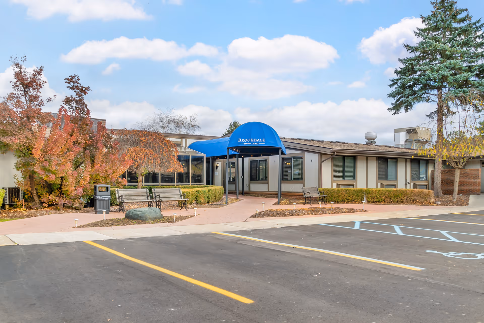 Exterior view of Brookdale Farmington Hills senior living facility with a blue awning over the entrance, benches along the walkway, autumn trees, and a parking lot in the foreground.