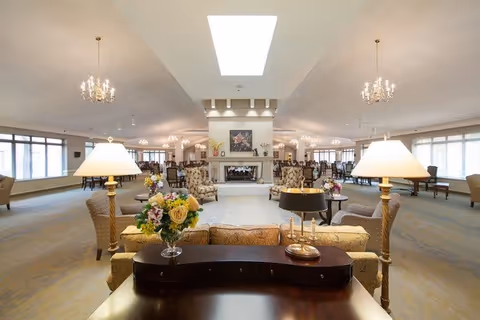 A spacious and well-lit living room area with large windows on both sides, featuring multiple seating arrangements including sofas and armchairs. There are two table lamps on a dark wooden table in the foreground with a vase of flowers and a candle holder. The room has a central fireplace with artwork above it, chandeliers hanging from the ceiling, and a skylight providing natural light.