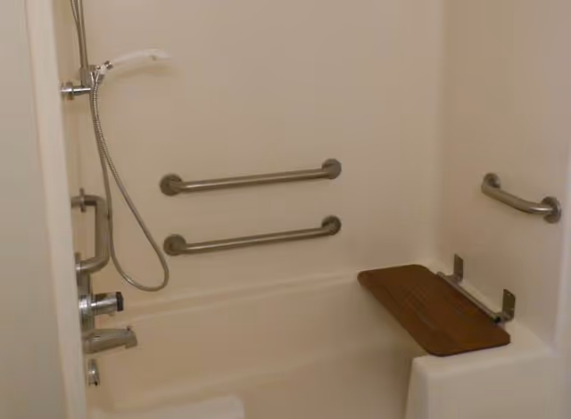 A bathtub with multiple stainless steel grab bars mounted on the walls for safety, a handheld showerhead attached to a flexible hose, and a foldable wooden shower seat installed on the right side of the tub.