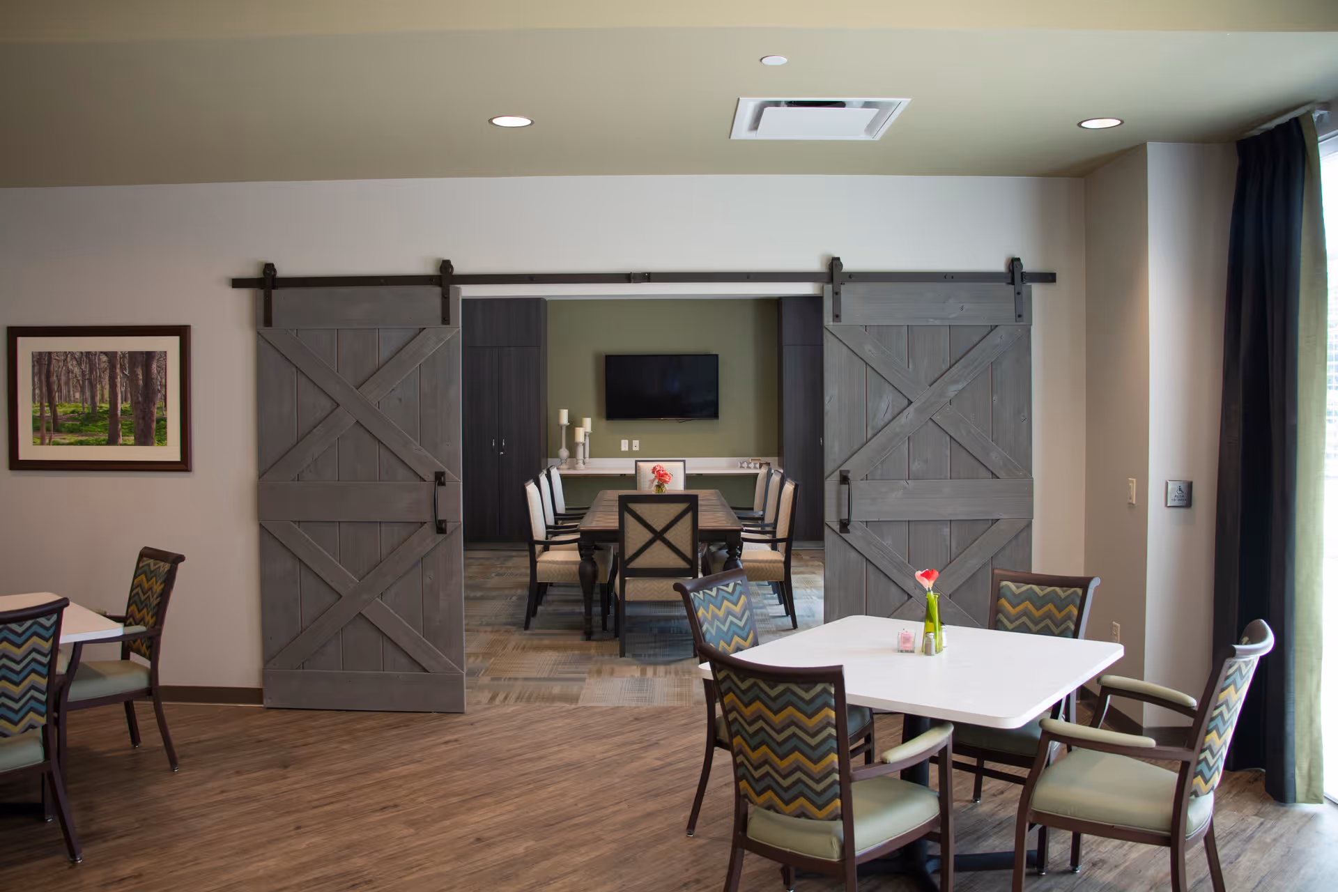 A dining area with a white table and four chairs with patterned upholstery. In the background, there is a room with a long wooden dining table surrounded by chairs, a wall-mounted TV, and decorative candles. The two rooms are separated by large sliding barn doors. The floor is wooden, and there is a framed picture on the wall to the left.