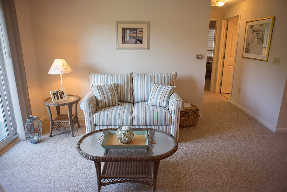Small living room with a striped loveseat, wicker coffee and side tables, a table lamp, and a hallway entrance.