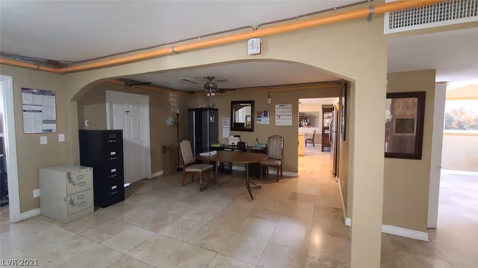 Interior view of a room with beige walls and tiled floor, featuring a small round table with two chairs, filing cabinets, a mirror on the wall, and a ceiling fan. The room opens into a kitchen area with visible appliances and dining furniture.