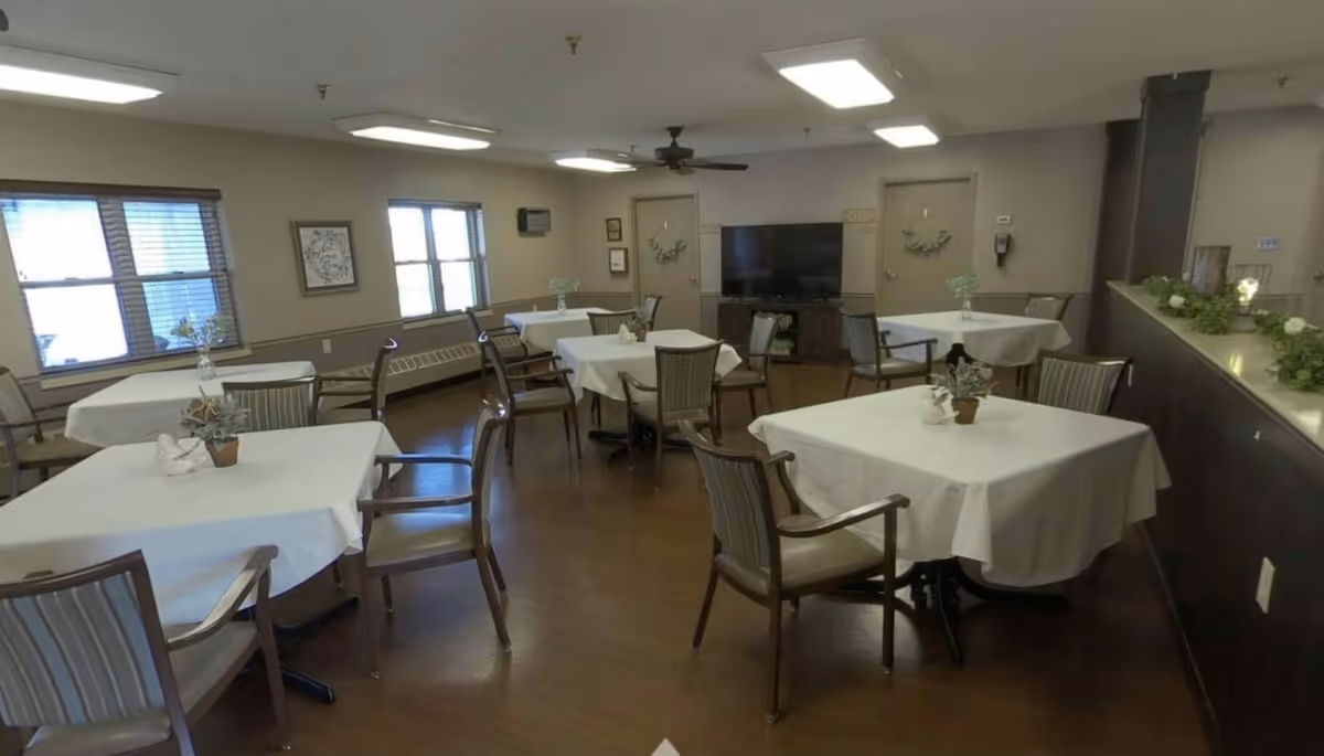 Dining room with multiple tables draped in white tablecloths, chairs, windows, and a TV on the far wall.