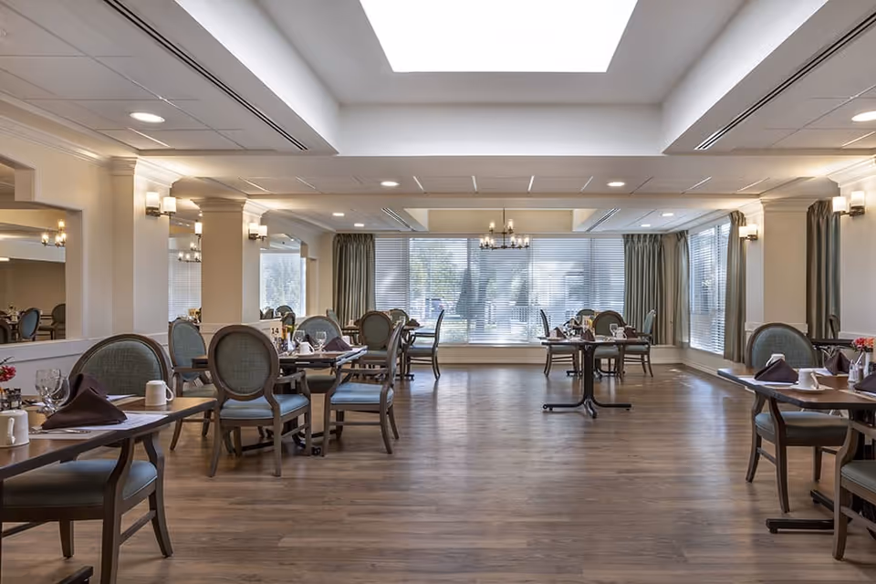 A spacious dining room with wooden floors, multiple tables set with plates, glasses, and napkins, green cushioned chairs, large windows with blinds and curtains, and ceiling lights including a chandelier.