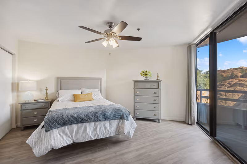 Bright bedroom with a bed, nightstand, dresser, ceiling fan, and sliding glass doors opening to a balcony view.