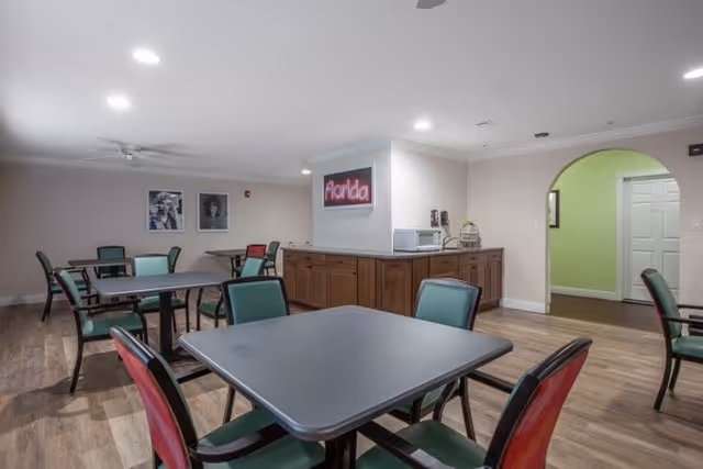 A dining area with several square tables and chairs with green and red backs. The room has wood flooring, beige walls, and ceiling lights. On one wall, there is a cabinet with a microwave and a neon sign that reads 'Florida'. There are two black and white framed pictures on the far wall and an arched doorway leading to another room with light green walls.