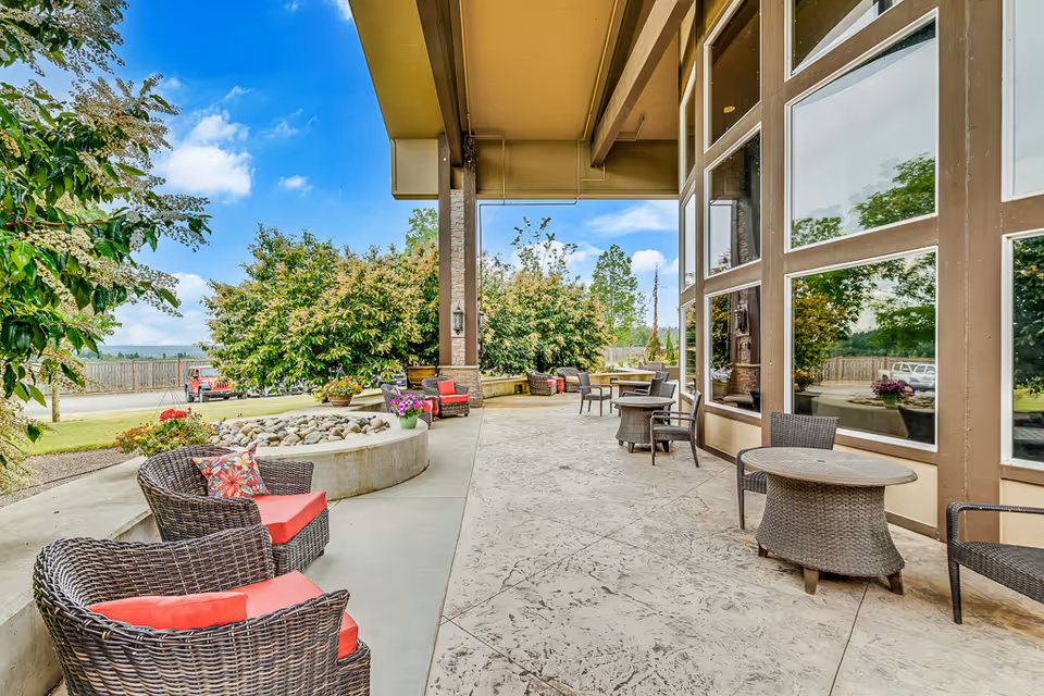 Outdoor covered patio area with wicker chairs and tables, some with red cushions and floral pillows. There are large windows reflecting greenery, potted plants, and trees in the background under a blue sky with some clouds.