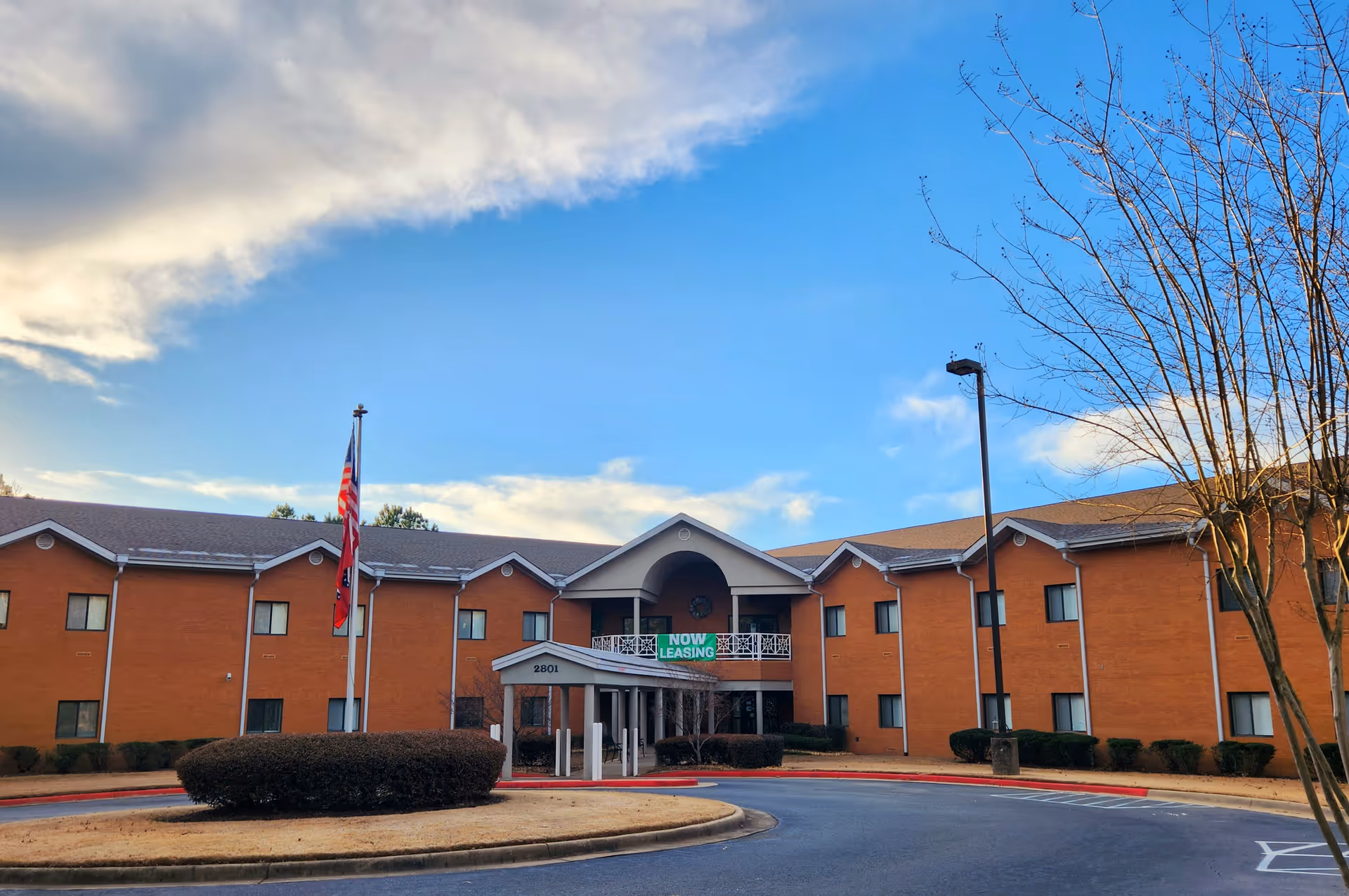 Front exterior of a two-story brick senior living building with a circular driveway, flagpoles, and a "Now Leasing" banner.