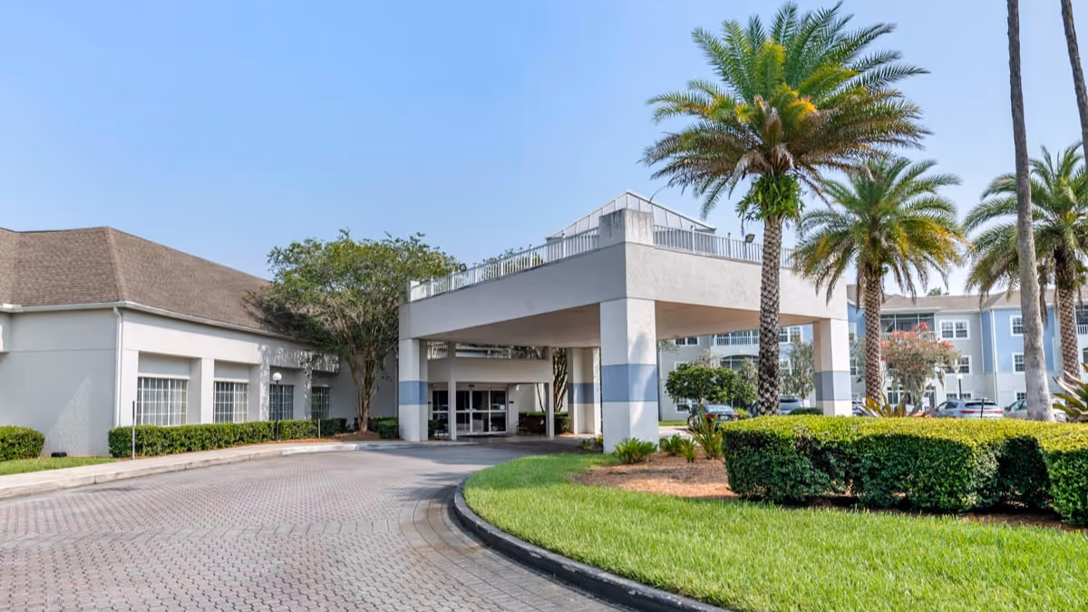 Exterior view of Brookdale Southside senior living facility showing the entrance with a covered drop-off area, palm trees, manicured bushes, and a clear blue sky.