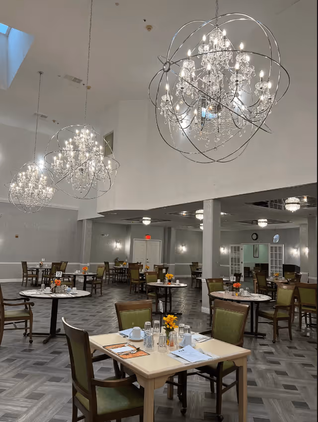 A spacious dining room with multiple tables and chairs arranged neatly. Each table is set with placemats, glasses, and small flower arrangements. The room features elegant chandeliers with crystal decorations hanging from the ceiling, and the floor has a patterned design. The walls are light-colored, and there are doors and windows visible in the background.