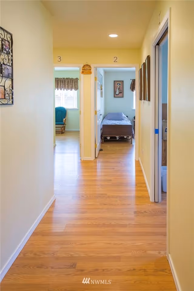 View down a hallway with light yellow walls and wooden flooring leading to two rooms numbered 3 and 2. Room 2 has a bed with a blue bedspread and a framed picture on the wall. Room 3 has a window with a curtain and a blue chair. There are picture frames on the right wall of the hallway.